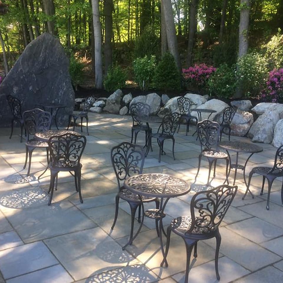 An outdoor patio with ornate black metal tables and chairs, surrounded by rocks, trees, and pink flowers.